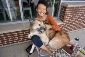 A smiling person sits outside on a chair with a large, fluffy dog on their lap. A glass table with a water bottle, a journal, and other items is in the foreground. Brick wall and window are in the background.