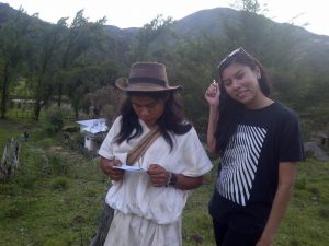 Two people stand outdoors on grassy terrain with mountains in the background. One, wearing traditional white clothing and a brown hat, reads a paper. The other, in a black graphic t-shirt and sunglasses, smiles and gestures with her hand.