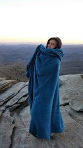 A person wrapped in a large blue blanket stands on a rocky cliff edge, overlooking a vast landscape of rolling hills at sunset.