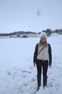 A woman wearing a white hat, cream sweater, black pants, and boots stands in a snowy field. Snow is falling, and a clump of snow appears to be mid-air above her head. Trees and shrubs are visible in the background.