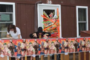 Three people stand on a porch decorated with a WWE-themed banner. One person is taped to a door with colorful tape, another dressed in black makes a playful pose, and a third person looks down at the railing.