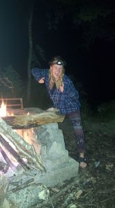 A person wearing a headlamp, plaid shirt, and patterned pants poses energetically beside a campfire at night in a wooded area, smiling and giving two thumbs up.