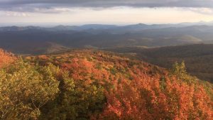 A scenic view of rolling mountains under a cloudy sky, with trees in the foreground displaying vibrant autumn colors of red, orange, and yellow. The distant hills fade into layers of blue and purple.