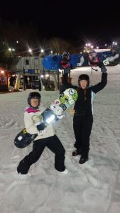 Two people in winter gear stand on snow at night, each holding a snowboard upright. They appear happy and excited, with a lit ski lodge and other people in the background.