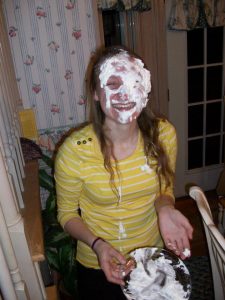 A person wearing a yellow striped shirt stands indoors, smiling with their face covered in whipped cream, holding an empty pie plate and gesturing playfully with their other hand.