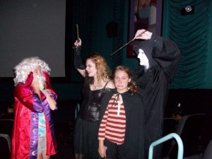 Four people in costume stand in a theater. One wears a white wig and red robe, another is in black with a wand, a third is in a striped shirt and cloak looking worried, and the fourth wears a white mask and black cloak, raising a wand.
