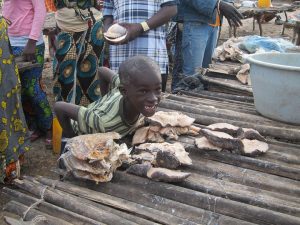 A smiling boy leans over a wooden table displaying dried fish at an outdoor market. Several adults stand nearby, and there is a large plastic basin on the right side of the image.