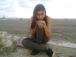 A young woman with long dark hair sits on a log at a beach, leaning forward with her arms resting on her knees and looking at the camera. The beach is mostly empty, with cloudy skies overhead.