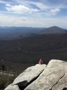 A person in a red plaid shirt sits on a large rocky outcrop overlooking a vast landscape of rolling, forested mountains under a blue sky with scattered clouds.