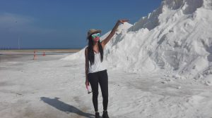 A woman wearing sunglasses, a hat, and casual clothes stands on a salt flat, smiling and raising one arm with a large mound of white salt in the background under a clear blue sky.