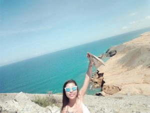 A woman wearing sunglasses smiles and raises her hat while standing on a rocky cliff overlooking turquoise ocean waters and a sandy, rugged coastline under a clear blue sky.