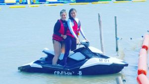 Two women wearing life jackets are sitting on a black and white jet ski in the water, smiling at the camera. There are floating barriers and water activity equipment in the background.