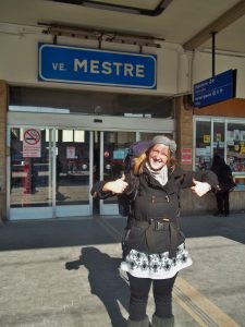 A smiling woman wearing a backpack and winter clothes stands outside the Venezia Mestre train station, pointing enthusiastically at the station sign above her.