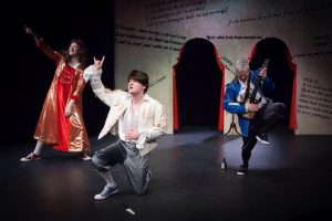 Three actors in theatrical costumes perform energetically on stage; one in a red dress, another kneeling in a white shirt, and the third playing a guitar in a blue jacket. A backdrop with text and two archways is behind them.