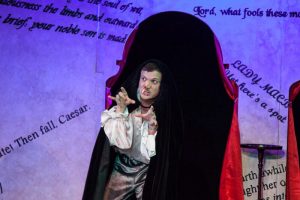 A man in theatrical costume and black cloak makes a dramatic, claw-like gesture on stage; the background features large, slanted text from classic literature on a light wall.