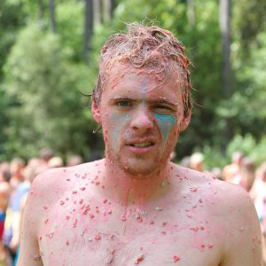 A man with messy hair and colorful paint and bits of food on his face and body stands outdoors, squinting and making a grimacing expression. Trees and a blurred crowd are visible in the background.