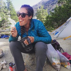A woman in sunglasses and a blue jacket sits on the ground at a campsite, smiling as she eats food. Behind her are trees, a tent, a mountain, and camping gear. The date 08/10/2015 is shown in the corner.