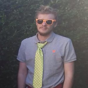 A man with light hair and a short beard wears orange sunglasses, a grey polo shirt, and a large, light green polka-dot tie. He stands outdoors in front of leafy greenery, looking at the camera.