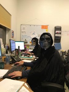 Two people wearing black cloaks and skull masks sit at desks with computers in an office. Papers, mugs, and monitors are visible, and a whiteboard covered with notes hangs on the back wall.