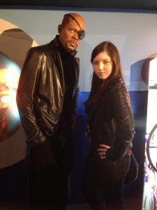 A woman posing next to a lifelike wax figure of Nick Fury, a character from Marvel comics, wearing a black eyepatch and black leather jacket, in what appears to be a museum or exhibit.