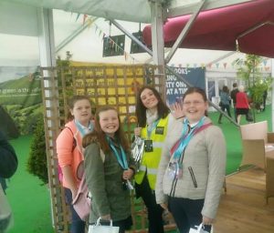 Four smiling girls stand together indoors at an event. One girl in a yellow vest waves at the camera. They all have lanyards and event passes around their necks. There are decorations and furniture in the background.