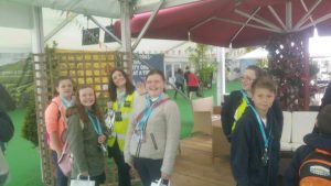 A group of smiling children and two adults wearing high-visibility vests stand together indoors under a canopy, some waving and holding bags, with green flooring and decorated seating in the background.