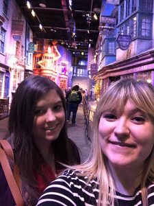 Two women smiling for a selfie inside a themed attraction resembling a magical, old-fashioned street with colorful, whimsical shopfronts and signs, reminiscent of Diagon Alley from the Harry Potter series.