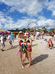 Two women in summer clothes stand on sandy ground at a lively outdoor festival, smiling and holding a hula hoop and pool noodles. Other people, a ferris wheel, and colorful tents are visible in the background under a blue sky.