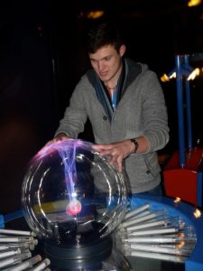 A person in a gray sweater places their hands on a large plasma globe, causing purple and pink electric streams to follow their touch inside the glass sphere. The background is dimly lit.