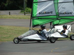 Two people wearing helmets ride side-by-side in small, three-wheeled sail-powered vehicles called blokarts on a paved track, with grass and pavement visible in the background.