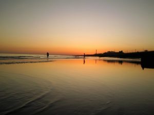 A tranquil beach at sunset with orange and purple hues in the sky, two silhouetted people near the shoreline, and gentle waves reflected in the wet sand.