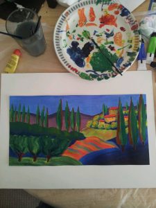 A colorful landscape painting on white paper shows trees, hills, and a house. Nearby are a paint palette with brushes, a cup of water, a yellow highlighter, and markers on a cluttered table.