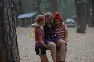 Three young women outdoors in a wooded area are laughing. Two, wearing red bandanas, lift the third, who is smiling and has a yellow bandana on her arm. Tents and a car are visible in the background.