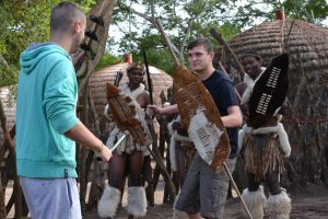 Two men in modern clothing interact with three people dressed in traditional Zulu attire, holding shields and spears, with round thatched huts and trees in the background.