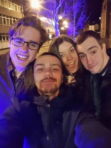 Four young adults—three men and one woman—pose closely together for a selfie on a city street at night, smiling under bright blue and yellow streetlights with trees and buildings in the background.