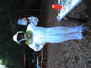 A person wearing a white beekeeping suit and veil stands outdoors on wood chips, holding up a clear container. There is a table with various items and red trees in the background.