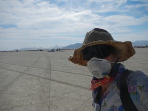 A person wearing a straw hat, sunglasses, a mask, and a colorful scarf stands on a flat, sandy desert landscape with mountains and scattered structures visible in the distance under a partly cloudy sky.