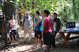 A group of boys stand outdoors in a wooded area, gathered around a person in a chef’s hat and apron. A wooden table with supplies is nearby, and sunlight filters through the trees.
