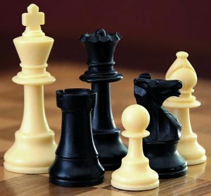 A close-up of chess pieces on a wooden chessboard, featuring a white king and pawn, and black queen, bishop, rook, knight, and pawn, with a blurred dark background.