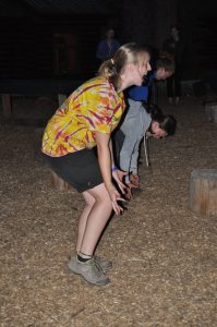 A group of young people outdoors at night, bending at the knees and leaning forward as if ready to jump, on a wood-chip covered ground surrounded by wooden stumps.