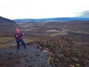 A person in outdoor gear stands on rocky terrain, giving two thumbs up, with a vast volcanic landscape and distant hills stretching out under a cloudy sky.