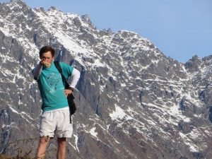 A person in a turquoise shirt and white shorts stands in front of a snowy mountain range, pointing toward the camera with one hand and resting the other on their hip. The sky is clear and blue.