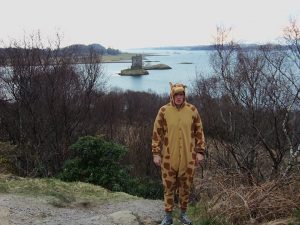 A person in a giraffe costume stands on a dirt path surrounded by leafless bushes, with a lake and a small stone building on an island visible in the background.