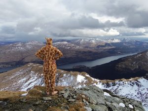 A person in a giraffe costume stands on a snowy mountain ridge, looking out over a scenic landscape of snow-capped peaks and a winding lake under a cloudy sky.