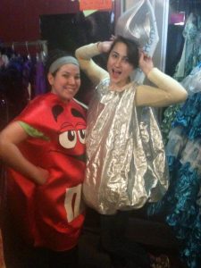 Two people smile while wearing costumes; one is dressed as a red M&M with a cartoon face, and the other is dressed as a shiny silver Hershey’s Kiss, posing with their arms raised playfully in a room filled with clothes.