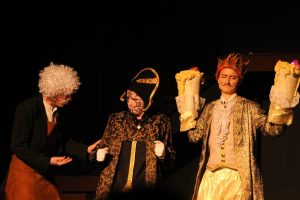 Three people in elaborate theatrical costumes perform on stage. One has a white wig and brown apron, another wears a black and gold outfit with face paint, and the third is dressed in gold with large gloves and a crown-like headpiece.