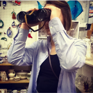 A person in a blue shirt looks through binoculars inside a shop filled with various ceramics and decorative items displayed on shelves and a pegboard wall.