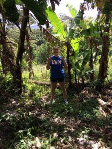 A person in a blue LVN shirt stands in a sunlit, tropical area surrounded by tall banana plants and lush greenery, with sunlight filtering through the leaves.