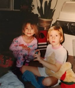 Two young children sit on the floor indoors, one in pink pajamas smiling at the camera, the other in a white T-shirt and blue socks holding a teddy bear and looking surprised. Toys and a potted plant are in the background.