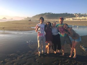 Five friends stand together smiling on a sandy beach at sunset, with gentle waves, distant hills, and buildings in the background. Two people wear tie-dye shirts, and everyone looks happy and relaxed.